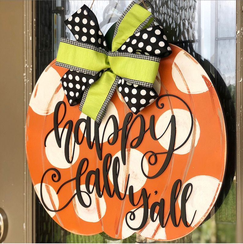 May include: Orange pumpkin-shaped door hanger with white polka dots and the text "happy fall y'all" in black script. A green and black polka dot bow is attached to the top.