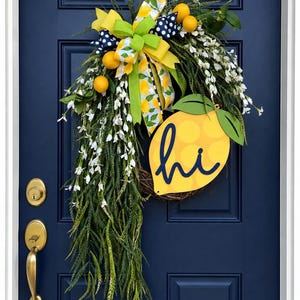 May include: A decorative wreath featuring a grapevine base, long green foliage, white floral sprigs, and yellow lemon accents. A large multi-patterned bow sits at the top, and a yellow lemon-shaped sign with the word hi in blue script is attached.
