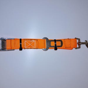 Op de afbeelding: Oranje veiligheidsgordel met een metalen gesp en een zilveren haak. De gordel is gemaakt van nylon webbing en heeft een zwarte band.