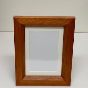 May include: A simple, rectangular picture frame made of light brown wood. The frame has a white mount and is ready to display a photo or artwork.