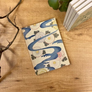 Puede incluir: Un pequeño cuaderno azul y blanco con un patrón floral y un diseño de onda azul. El cuaderno está sobre una superficie de madera con una rama y una planta verde en el fondo.