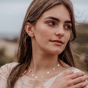 May include: A delicate silver necklace featuring small, round disc charms. The necklace is worn by a person with long, light brown hair and a neutral-toned top. The background is blurred, suggesting an outdoor setting.