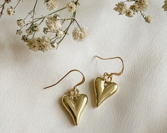 Può includere: Un paio di orecchini pendenti a forma di cuore dorati con un delicato design a filo. Gli orecchini sono sospesi da un semplice gancio.