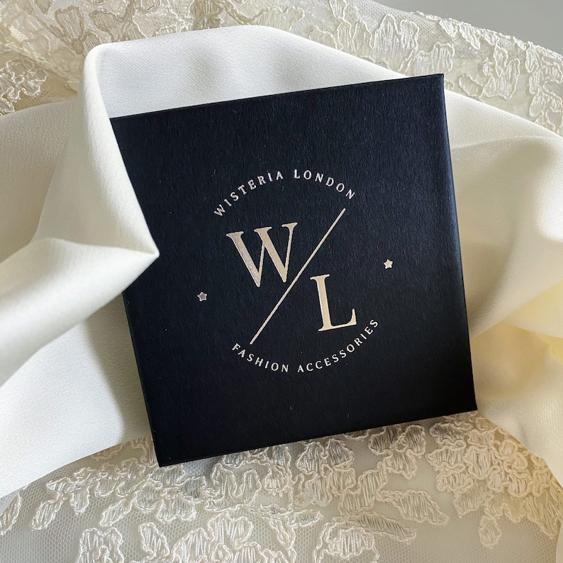 K&ouml;nnte beinhalten: Eine schwarze Box mit einem wei&szlig;en und goldenen Logo, auf dem "Wisteria London Fashion Accessories" steht, mit einem stilisierten "W" und "L", die sich &uuml;berlappen.