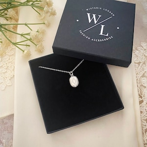 Może przedstawiać: Srebrny naszyjnik z małym owalnym wisiorkiem w czarnym pudełku. Pudełko ma nadrukowany tekst "Wisteria London" i "Fashion Accessories".