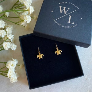 Peut inclure: Une paire de boucles d'oreilles en forme de tortue dorées dans un coffret à bijoux noir. Le coffret porte l'inscription "Wisteria London Fashion Accessories" sur le dessus.