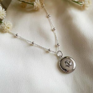May include: A silver chain necklace with a small silver locket featuring a bee design. The necklace is on a white fabric background.