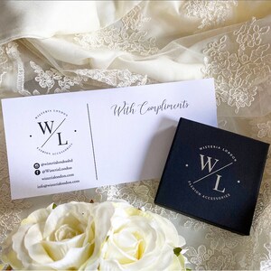 K&ouml;nnte beinhalten: Eine schwarze Geschenkbox mit einem wei&szlig;en Logo, auf dem "WISTERIA LONDON" und "W/L FASHION ACCESSORIES" steht. Die Box steht auf einem wei&szlig;en Spitzenstoff mit einer wei&szlig;en Karte, auf der "Mit freundlichen Gr&uuml;&szlig;en" steht und ein Logo mit der Aufschrift "WISTERIA LONDON" und "W/L FASHION ACCESSORIES" zu sehen ist.
