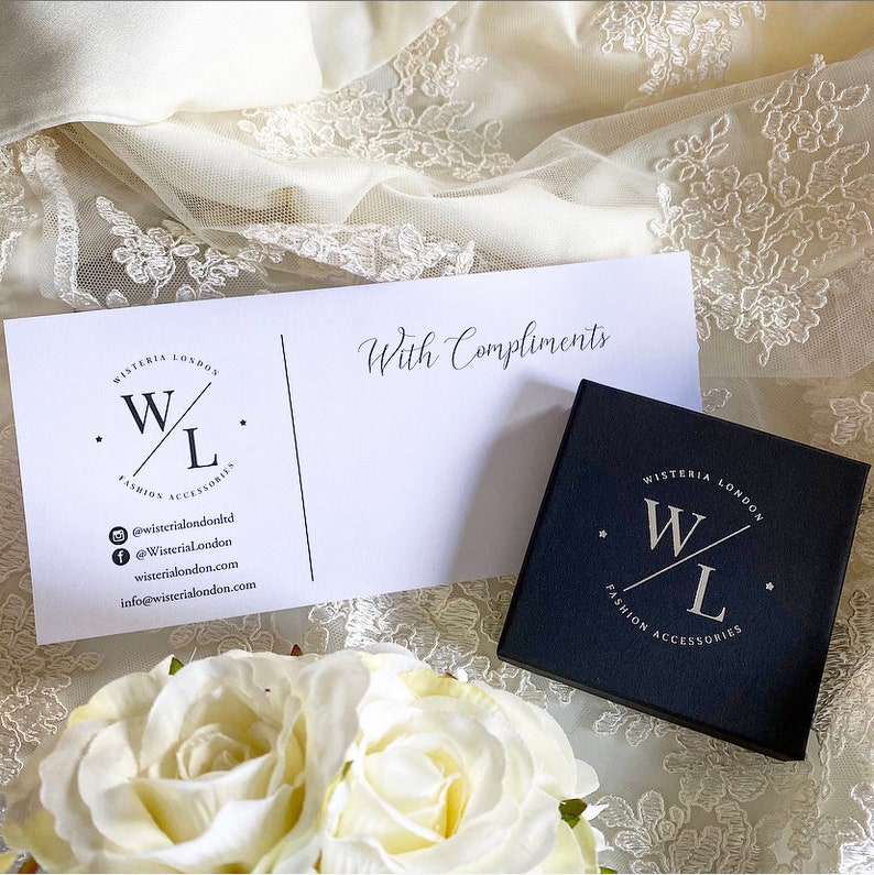K&ouml;nnte beinhalten: Eine schwarze Geschenkbox mit einem wei&szlig;en Logo, auf dem "WISTERIA LONDON" und "W/L FASHION ACCESSORIES" steht. Die Box steht auf einem wei&szlig;en Spitzenstoff mit einer wei&szlig;en Karte, auf der "Mit freundlichen Gr&uuml;&szlig;en" steht und ein Logo mit der Aufschrift "WISTERIA LONDON" und "W/L FASHION ACCESSORIES" zu sehen ist.