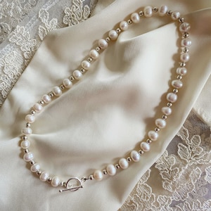 May include: A white pearl necklace with a silver toggle clasp. The necklace is on a white satin fabric background.