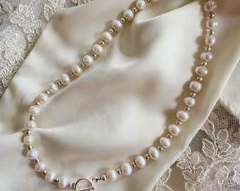 May include: A white pearl necklace with a silver toggle clasp. The necklace is on a white satin fabric background.