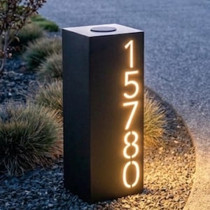 May include: A black, rectangular address sign with illuminated numbers 15780. The numbers glow with a warm, inviting light. The sign is set in a garden with small stones and greenery.