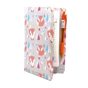 May include: A gray and white notebook cover with a repeating pattern of foxes, trees, and birds. The cover has a built-in pen holder with an orange pen.