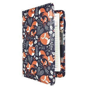 May include: A navy blue journal with a repeating pattern of orange foxes, white berries, and green sprigs. The journal is open, revealing lined pages and a pen. The pen and the inside cover also feature the fox pattern.