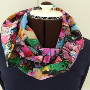 May include: An infinity scarf featuring a vibrant, abstract print with bold splashes of magenta, bright green, blue, and orange against a black background. The fabric is draped in multiple folds, showcasing a complex, multi-coloured pattern.