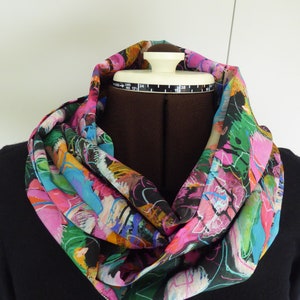 May include: A brightly coloured infinity scarf with an abstract pattern. The scarf displays pink, blue, green, and black hues. The scarf is draped around a mannequin. The mannequin has a measurement of 33 cm.