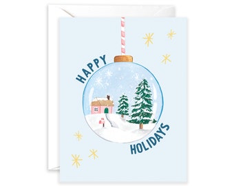 May include: A blue Christmas card with a white envelope. The card features a snow globe ornament with a pink house, two green trees, and a snowy landscape. The text "Happy Holidays" is written in blue on the card.