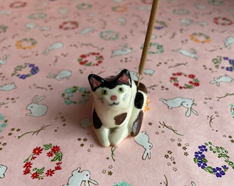 May include: A white and black ceramic cat incense holder with a brown nose and green eyes. The cat is sitting with its tail curled around its legs. A brown incense stick is sticking straight up from the cat's back.