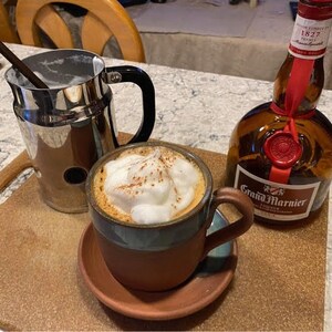 May include: A brown ceramic mug with a blue rim filled with a cappuccino topped with whipped cream and cinnamon. A silver pitcher with a handle and a bottle of Grand Marnier liqueur are also in the image.
