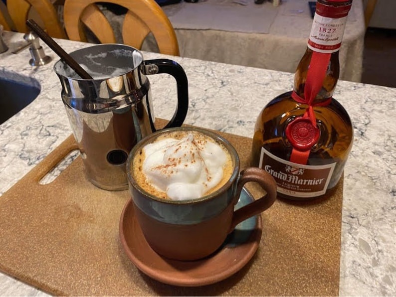 May include: A brown ceramic mug with a blue rim filled with a cappuccino topped with whipped cream and cinnamon. A silver pitcher with a handle and a bottle of Grand Marnier liqueur are also in the image.