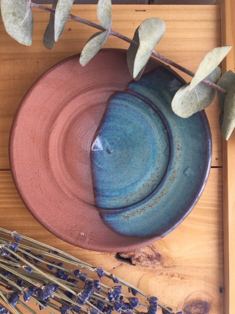 May include: A small, round ceramic dish with a brown and blue glaze. The dish is half brown and half blue, with a smooth, rounded edge.