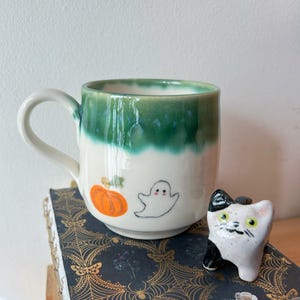 Könnte beinhalten: Eine weiße Keramiktasse mit grüner Glasur und Halloween-Motiv. Die Tasse ist mit einem Geist, einem Kürbis und Blättern bemalt. Eine kleine weiße Keramikkatzenfigur mit schwarzen Markierungen steht neben der Tasse.