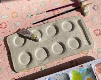 May include: A white ceramic paint palette with 10 round wells and a long, thin groove for a palette knife. The palette is speckled with gray flecks and has a tube of paint on top.