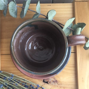 May include: A brown ceramic mug with a green rim and a brown saucer. The mug is empty and the saucer is on a wooden surface.