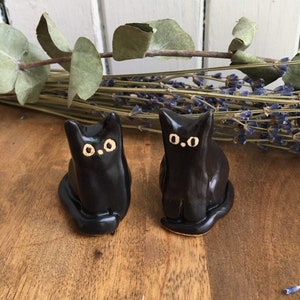 Puede incluir: Dos figuritas de gato de cerámica negra con ojos blancos. Los gatos están sentados con sus colas enroscadas alrededor de sus patas.