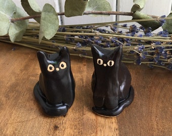 Puede incluir: Dos figuritas de gato de cerámica negra con ojos blancos. Los gatos están sentados con sus colas enroscadas alrededor de sus patas.