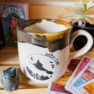 Puede incluir: Taza de cerámica blanca con un gráfico negro de "Witch Club" que presenta una bruja en una escoba, una luna creciente y un horizonte. La taza tiene un borde marrón y dorado y un asa blanca. Una pequeña figura de gato gris y cartas del tarot están cerca.