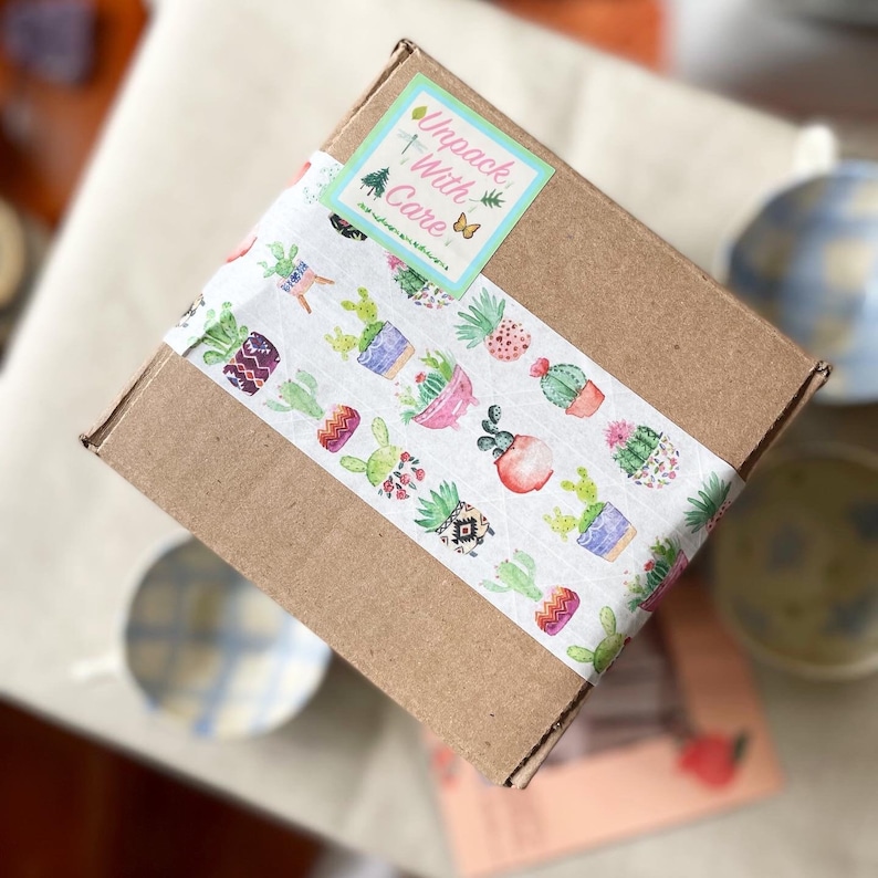 May include: A brown cardboard box with a white and green patterned tape. The tape features a variety of cactus plants in pots. The box has a pink and green sticker that says "Unpack With Care".