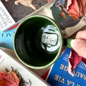 Könnte beinhalten: Eine grüne Keramiktasse mit den Worten "The End" im Inneren. Die Tasse ist von Büchern und Herbstblättern umgeben. Die Tasse wird von einer Person gehalten.