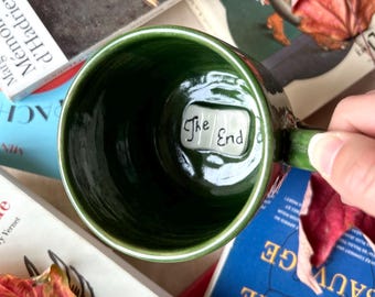 May include: A green ceramic mug with the words "The End" inside. The mug is surrounded by books and autumn leaves. The mug is being held by a person.
