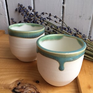 May include: Two white ceramic mugs with a teal glaze around the rim. The glaze has a drip effect on one of the mugs.