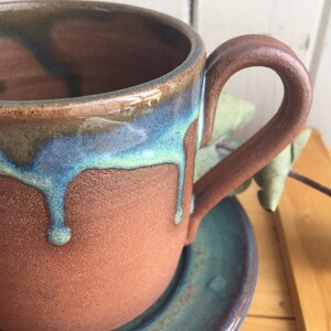 May include: A brown ceramic mug with a teal glaze dripping down the side. The mug has a rounded handle and sits on a matching saucer.