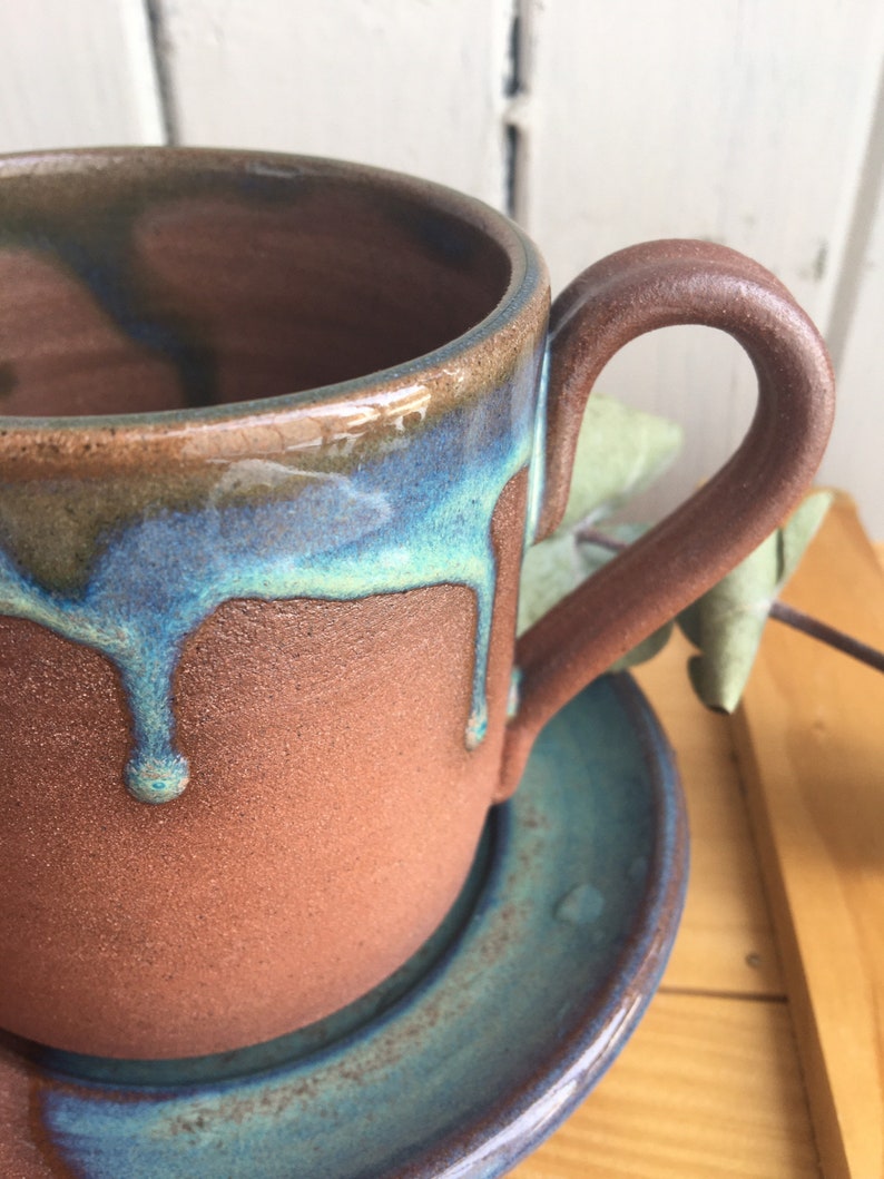 May include: A brown ceramic mug with a teal glaze dripping down the side. The mug has a rounded handle and sits on a matching saucer.