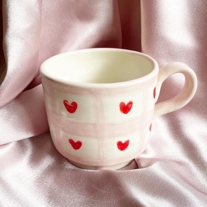 May include: A pink and white ceramic mug with a checkered pattern and red hearts. The mug has a rounded handle.