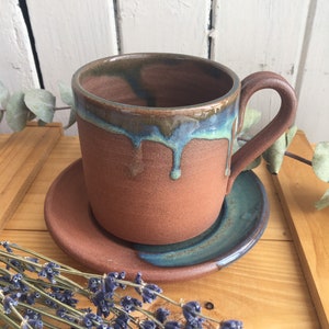 May include: A terracotta mug with a blue and green glaze, dripping down the side. The mug is sitting on a matching saucer.