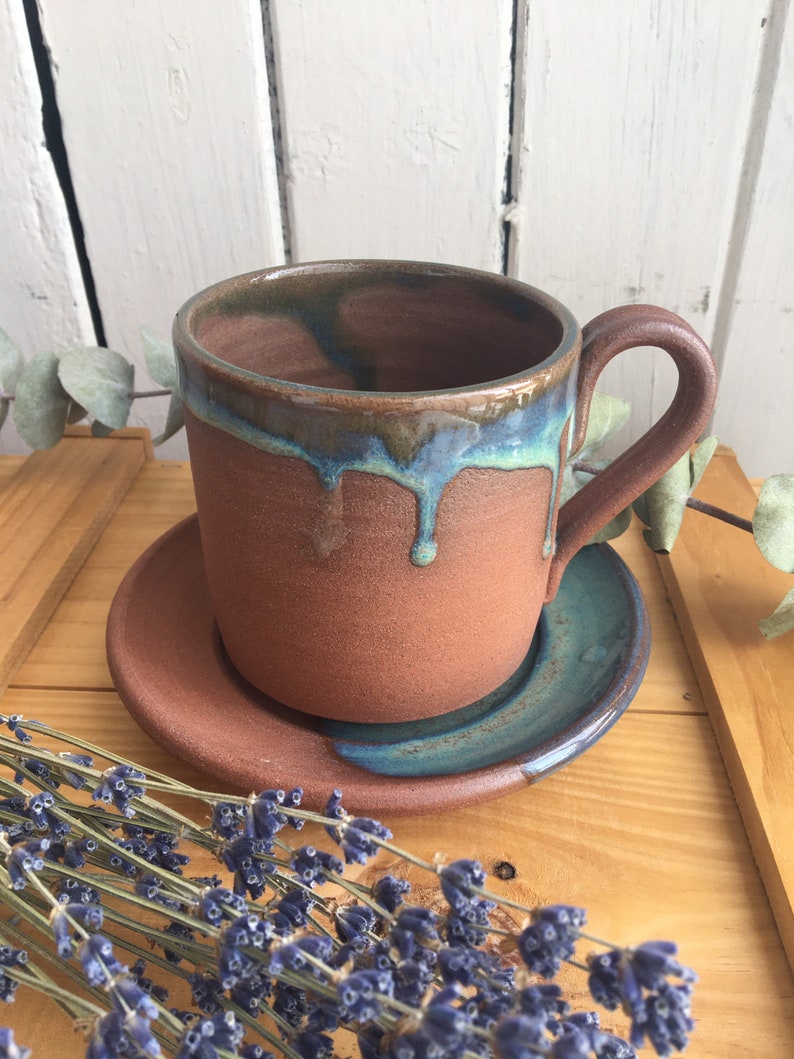 May include: A terracotta mug with a blue and green glaze, dripping down the side. The mug is sitting on a matching saucer.