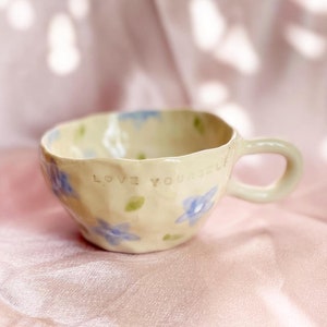 May include: A white ceramic mug with a blue floral design and the words "Love Yourself" written on the side.