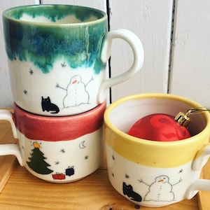 Op de afbeelding: Drie keramische mokken met winterse illustraties. De bovenste mok heeft een groenblauwe glazuur, de middelste is rood en de onderste is geel. Elke mok toont een sneeuwman, een zwarte kat en sneeuwvlokken.
