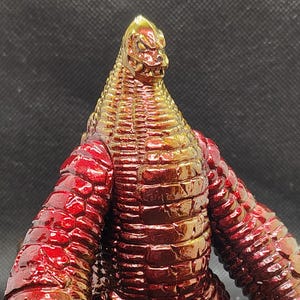 May include: A metallic red and gold monster figurine with a textured, scaly appearance. The figure has a reptilian head, large arms, and a stout body. The toy is posed with its arms at its sides and its feet planted firmly on the ground.