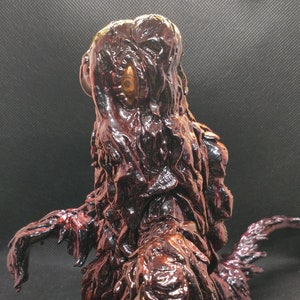 May include: A dark red and gold resin figurine of a creature with a textured, dripping surface. The creature has a long, pointed tail and a small, round head with a single, large eye.