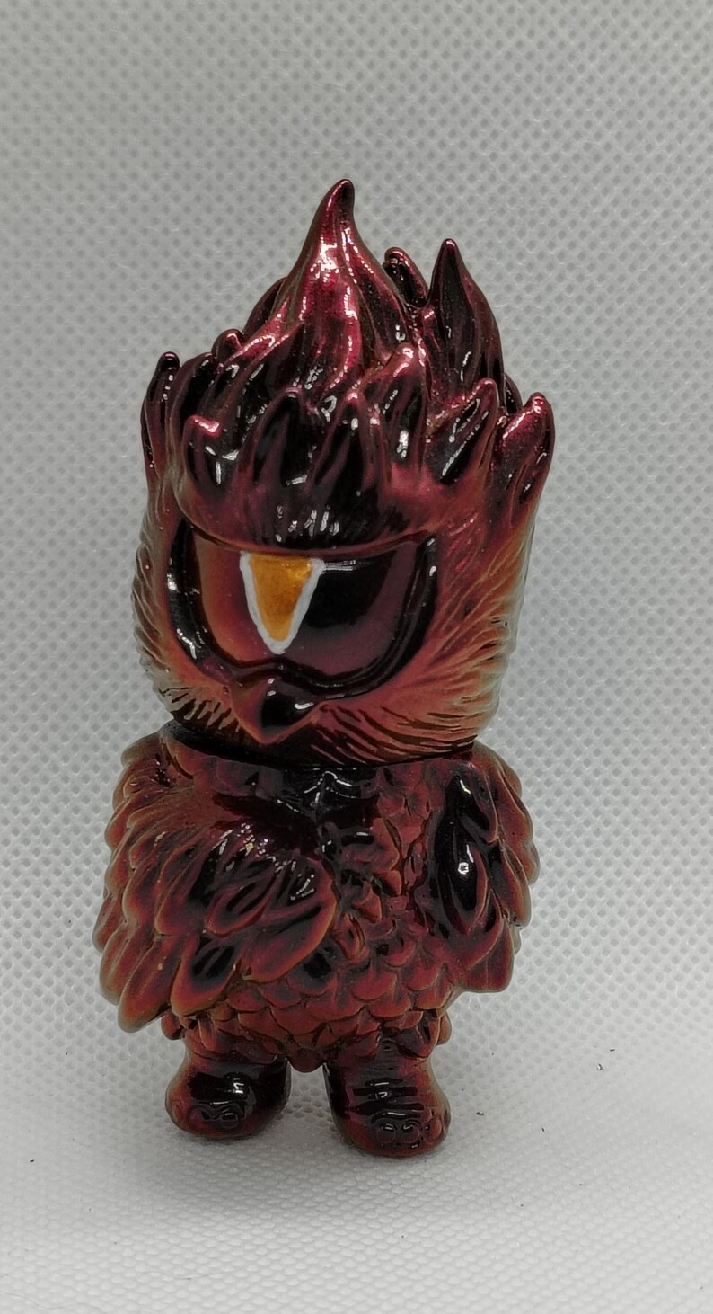 K&ouml;nnte beinhalten: Stilisierte Eulenfigur in Rot mit dunkleren Akzenten. Die Figur hat eine feurige, strukturierte Oberfl&auml;che. Das Design umfasst ein gro&szlig;es Auge mit gelb-wei&szlig;em Iris. Die Figur hat ein einzigartiges, k&uuml;nstlerisches Design.