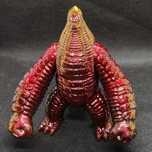 May include: A metallic red and gold figurine of a creature with a reptilian head and textured body. The figure has large arms and feet, and stands in a crouching position. The color transitions from gold to red.