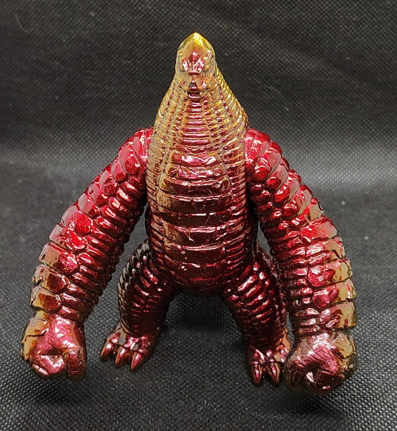 May include: A metallic red and gold figurine of a creature with a reptilian head and textured body. The figure has large arms and feet, and stands in a crouching position. The color transitions from gold to red.
