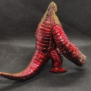May include: A metallic red and gold figurine of a reptilian creature with a textured, scaly surface. The figure has a long tail, large arms, and stands on two legs. The color transitions from gold at the head to red on the body.