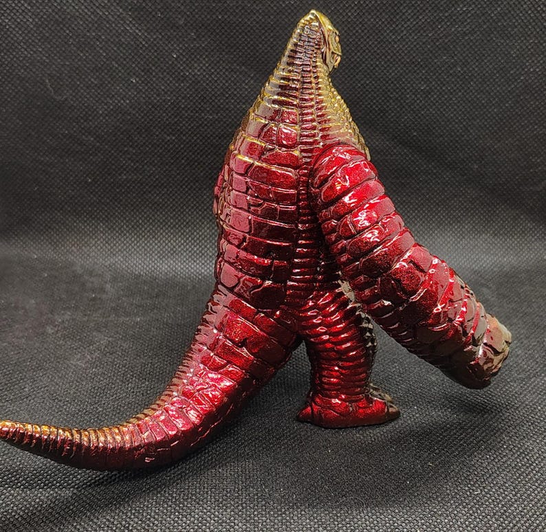 May include: A metallic red and gold figurine of a reptilian creature with a textured, scaly surface. The figure has a long tail, large arms, and stands on two legs. The color transitions from gold at the head to red on the body.