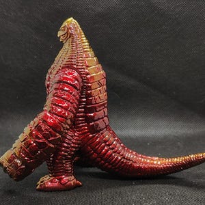 May include: A red and gold figurine of a reptilian creature with a long tail and textured skin. The figure has a detailed head and is posed in a dynamic stance, showcasing its intricate design and metallic finish.